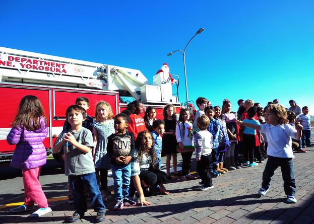 Kinder vor einer Feuerwache mit der Aufschrift 'Yokosuka Fire Department' auf einer Straße mit Laternenmästen, Bäumen und einem klaren blauen Himmel.