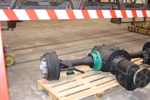 Ein Nahaufnahme einer Metallachse auf einer Holzpalette in einem Lagerhaus, umgeben von einer roten und weißen gestreiften Absperrung und mehreren anderen Metallobjekten im Hintergrund.