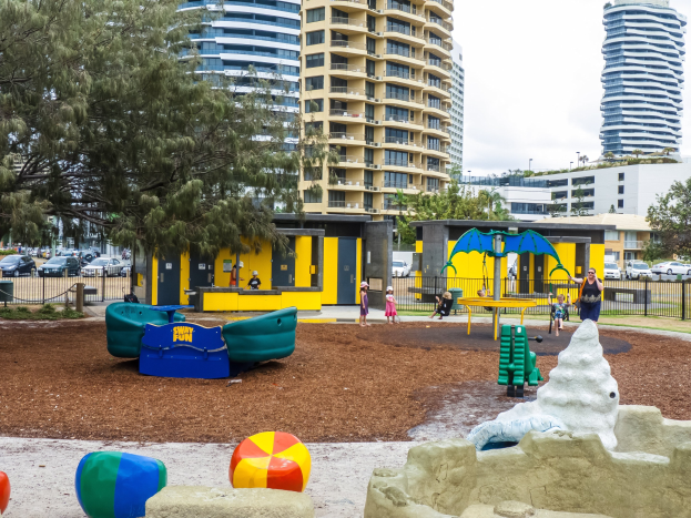 Spielplatz mit Kindern auf Geräten, umgeben von Bäumen und Stadtgebäuden, mit Fahrzeugen auf der Straße und Himmel darüber.