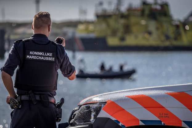 Ein Polizist in Uniform steht neben einem Polizeiwagen an einem Gewässer und hält ein Funkgerät in der Hand. Im Hintergrund ist ein Boot auf dem Wasser zu sehen, auf dem sich ein paar Menschen befinden, und der Himmel ist oben im Bild zu erkennen.
