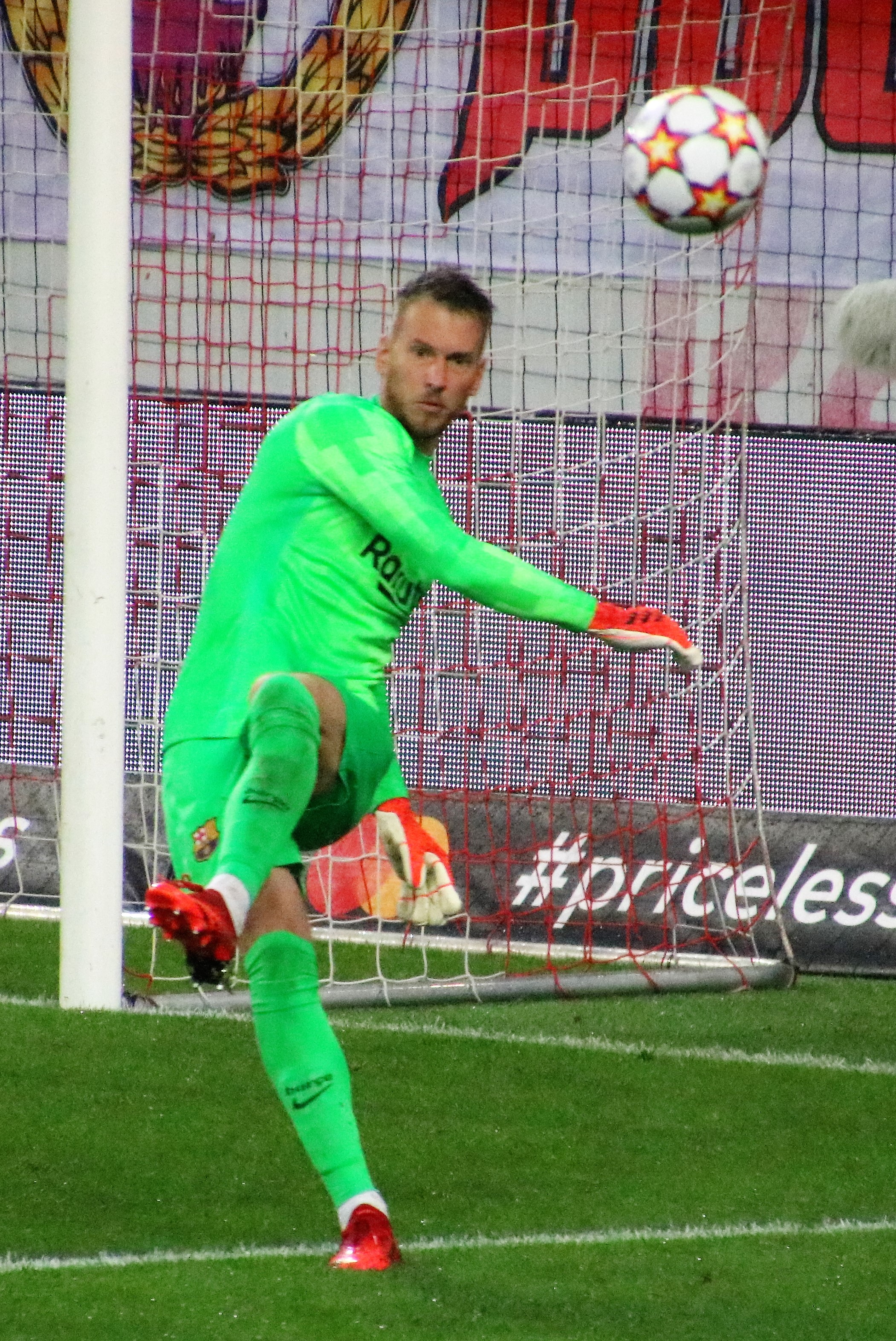 Ein Fußballspieler in grüner Uniform fängt einen Ball vor einem Tor mit Netz und einem Banner im Hintergrund, trägt Fußballhandschuhe.