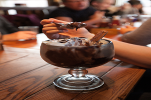 Eine Gruppe von Menschen sitzt um einen Tisch mit einem Glas Schokoladenpudding in der Mitte, eine Person im Vordergrund isst es mit einem Löffel, der Hintergrund ist unscharf.