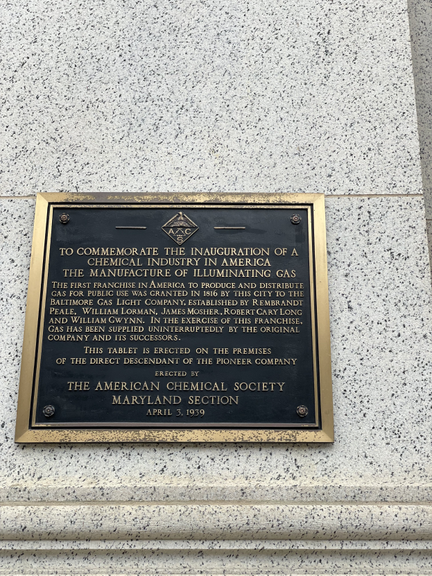 Eine Messingtafel an einer Gebäudewand mit fetter schwarzer Schrift, die "Zur Erinnerung an die Einweihung der Chemischen Industrie in Amerika" lautet, umgeben von einem dekorativen Rahmen.