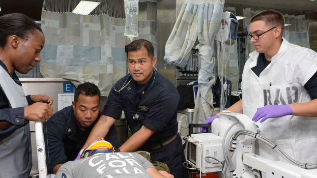 Eine Gruppe von vier Personen um einen Mann auf einer Trage in einem Krankenhauszimmer mit medizinischer Ausrüstung, Vorhängen, einer Kochsalzlösung und Deckenlampen, wahrscheinlich in einer Notaufnahme der US-Küstenwache.