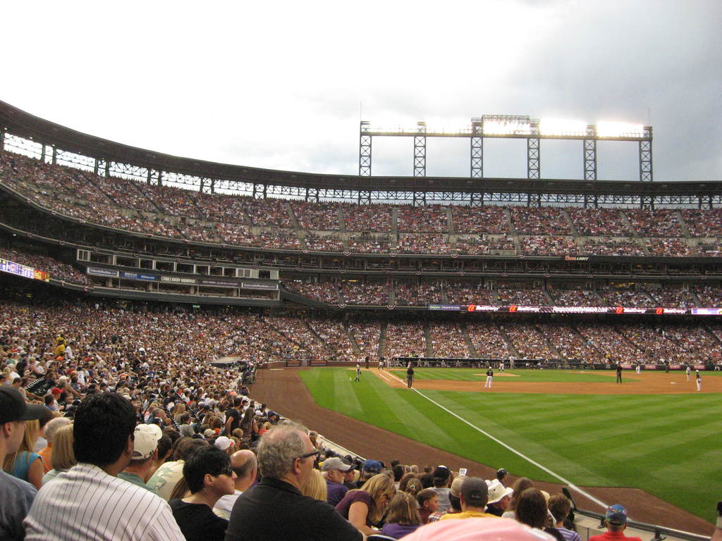 Ein Cricket-Stadion mit Spielern auf dem Feld und Zuschauern in den Rängen, unter einem Metallrahmen mit Scheinwerfern.