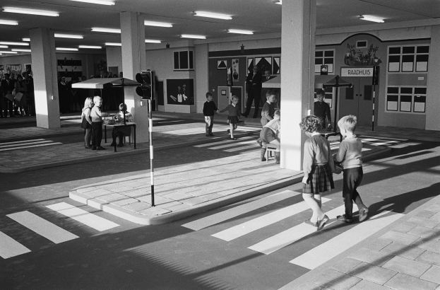 Schwarzes und weißes Foto von Kindern, die eine Straße an einer Fußgängerüberführung überqueren, mit Fußgängern in der Nähe, einem Verkehrssignalmast im Vordergrund und Säulen mit Deckenleuchten im Hintergrund.