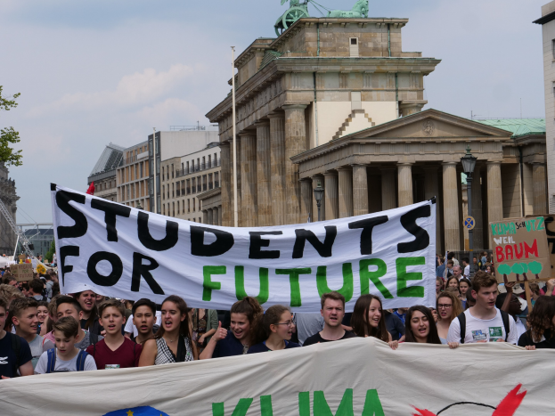 Gruppe von Schülern marschiert in Berlin mit einem bunt bemalten "Students for Future"-Schild gegen den Hintergrund von Gebäuden, Bäumen und Himmel.