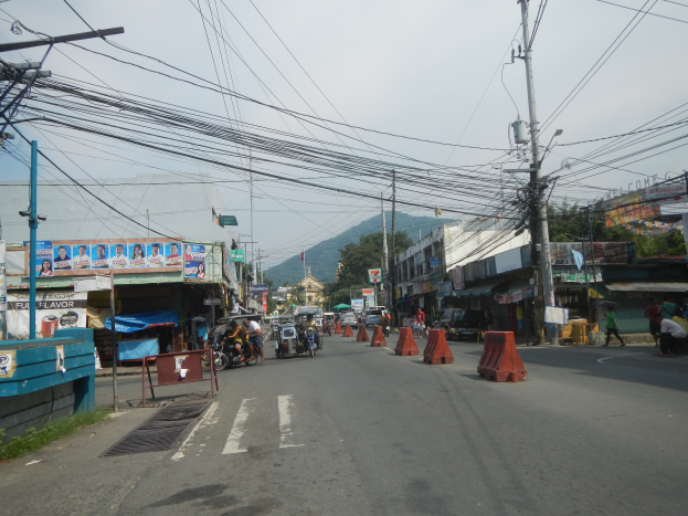 Eine Stadtstraßenszene mit Strommasten, Fahrzeugen, Fußgängern, Gebäuden, Werbetafeln, Bäumen, Hügeln und einem klaren blauen Himmel.