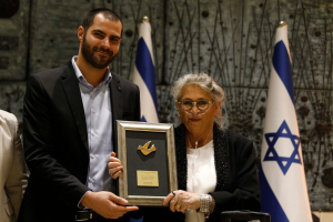 Ein Mann und eine Frau stehen nebeneinander und halten jeweils einen Preis in der Hand, wobei der Mann ein schwarzes Sakko, ein weißes Hemd und schwarze Hosen trägt und die Frau eine Brille trägt, vor einem Hintergrund aus blauen, weißen und cremefarbenen Flaggen und einer Wand.