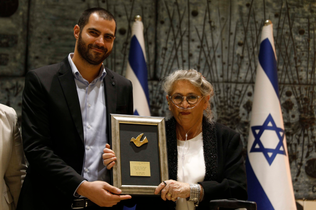 Ein Mann und eine Frau stehen nebeneinander und halten jeweils einen Preis in der Hand, wobei der Mann ein schwarzes Sakko, ein weißes Hemd und schwarze Hosen trägt und die Frau eine Brille trägt, vor einem Hintergrund aus blauen, weißen und cremefarbenen Flaggen und einer Wand.