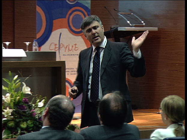 Ein Mann in einem schwarzen Blazer steht an einem Podium mit Mikrofonen, spricht in ein tragbares Mikrofon, mit einer Blumendekoration und einem Tisch mit einem Buch und einer Wasserflasche in der Nähe, während drei Personen in Stühlen vor ihm sitzen, vor einem Hintergrund aus einer braunen Wand und einem bunten Brett mit Text.