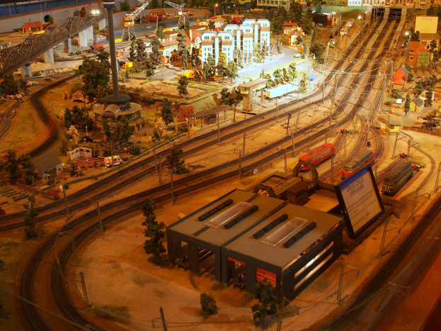 Eine detaillierte Modellbahnausstellung in einem Museum, mit einer Miniaturstadtlandschaft mit Gebäuden, Bäumen, Schienen, Fahrzeugen, Pfählen, Lichtern und anderen Gegenständen.