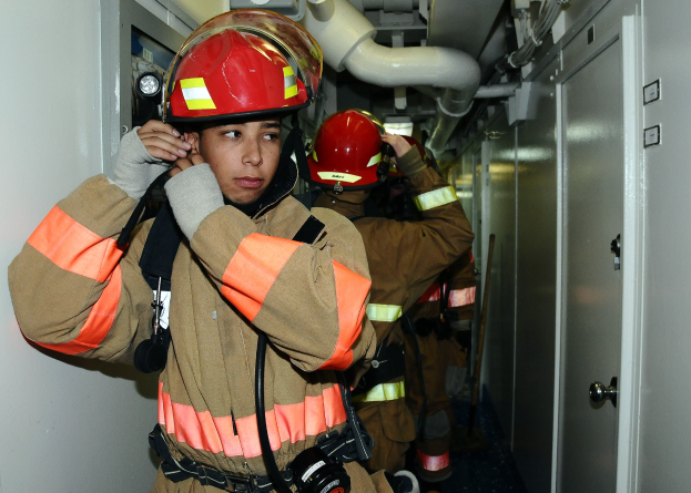 Gruppe von Feuerwehrleuten in Uniform, die in einem Raum mit Rohren und Gegenständen im Hintergrund stehen, wahrscheinlich während einer Übung.