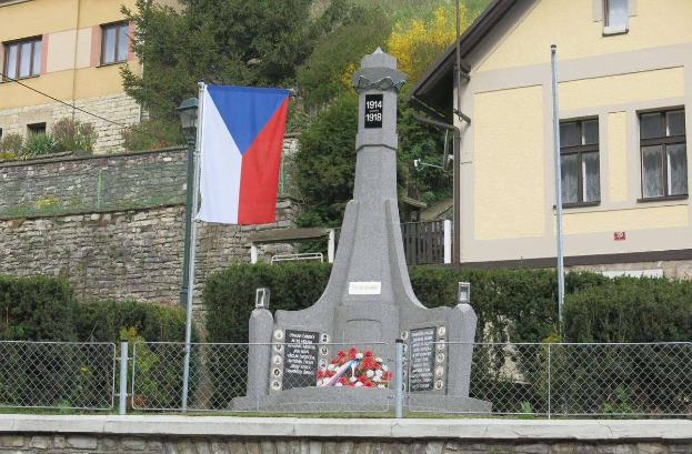 Ein Denkmal mit einer Fahne oben drauf, umgeben von einem Zaun, Pflanzen, Bäumen und einer Wand, vor einem Gebäude, gewidmet denen, die ihr Leben während des Zweiten Weltkriegs verloren haben.