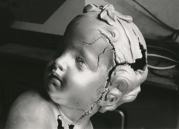 Schwarzes und weißes Foto einer Skulptur, die ein Kind mit gebrochenem Kopf zeigt, umgeben von Drähten und einer Wand im Hintergrund.