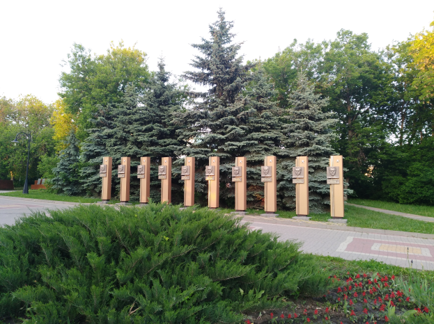 Eine Parkszene mit einem Gedenktafel für Chernobyl-Opfer, umgeben von Bäumen, Blumen, Gras, einem Weg und einem Straßenmast unter einem bewölkten Himmel.