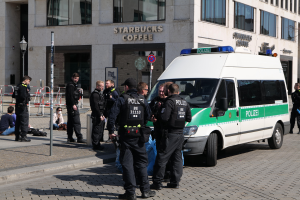 Eine Gruppe von Polizisten steht vor einem Starbucks-Café, rechts daneben ein Van geparkt, links einige Menschen, im Hintergrund ein Gebäude mit Fenstern, einer Schautafel, einem Laternenmast und einem Zaun.