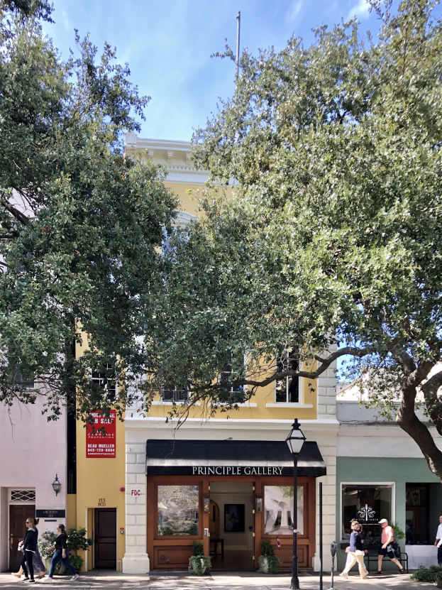 Außenansicht der Principle Gallery in Charleston, SC, mit Menschen auf dem Bürgersteig, Topfpflanzen, einer Straßenlaterne, einem Schild, einer Fahne, einem Flaggenmast und einem bewölkten Himmel.
