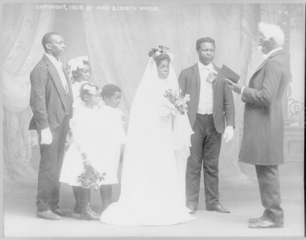 Ein Schwarz-Weiß-Foto einer Braut in einem weißen Kleid und Schleier, die einen Strauß hält und neben einem Bräutigam mit einem Buch steht, umgeben von ihrer Familie und Vorhängen im Hintergrund.