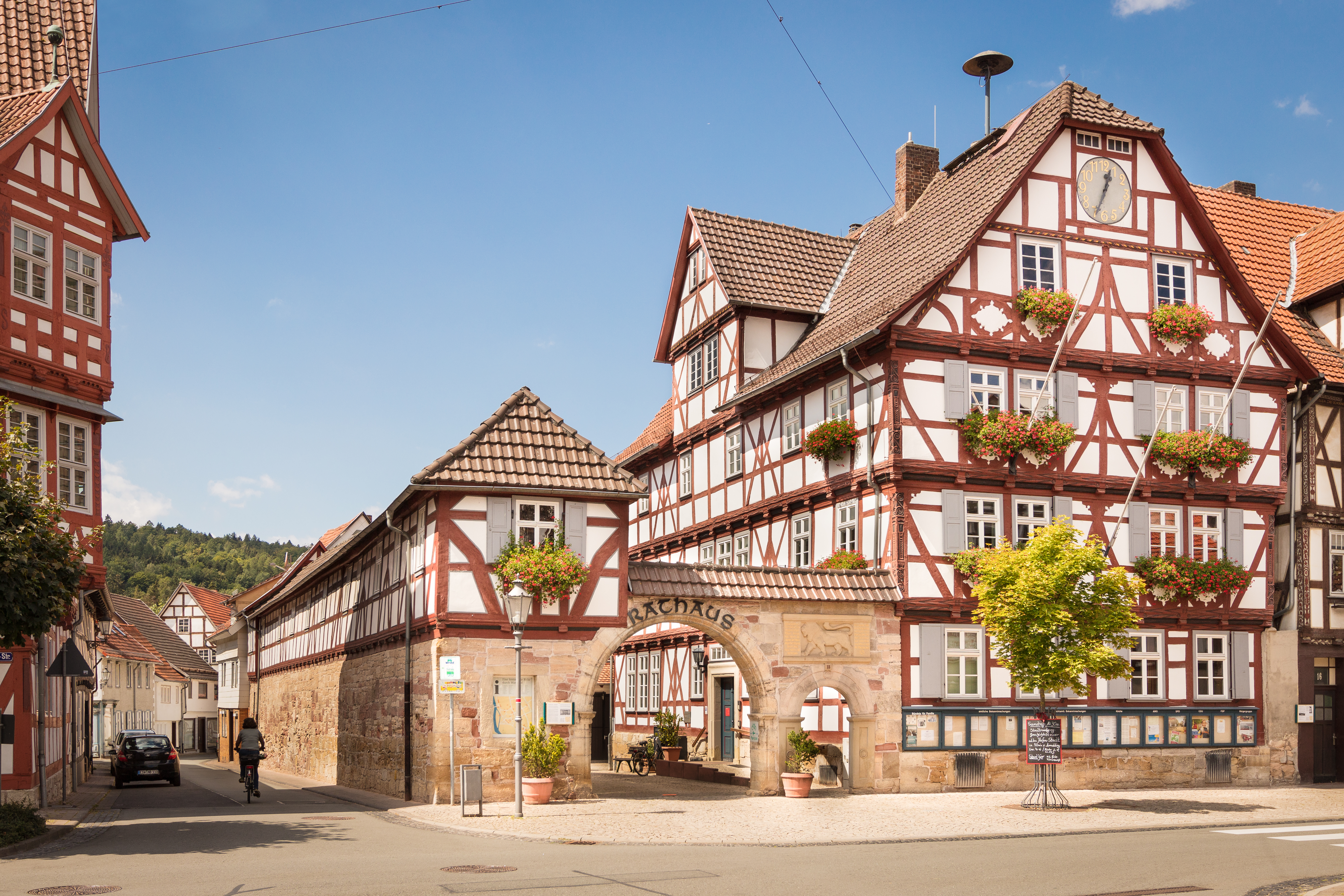 Eine Straße in Rothenburg ob der Tauber, Deutschland, mit alten Gebäuden mit Fenstern, Bäumen, Laternen und einer Person auf einem Fahrrad, mit Hügeln und einem bewölkten Himmel im Hintergrund.