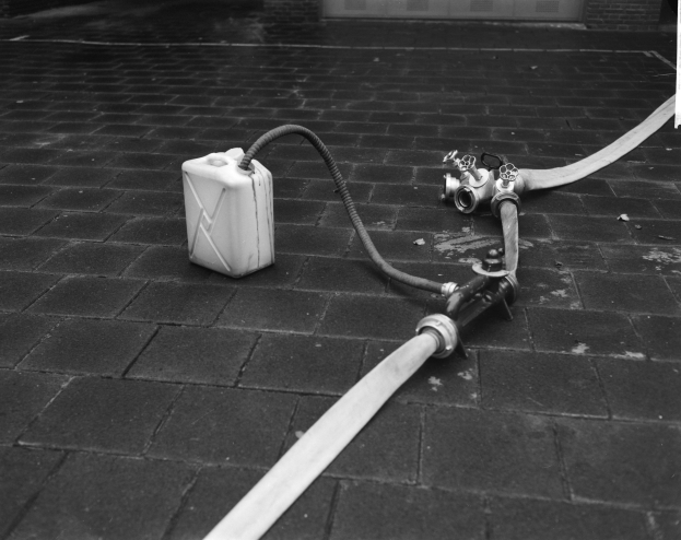 Schwarz-weißes Foto eines Gasbehälters mit einem Schlauch, der vor einem Gebäude mit einer Tür im Hintergrund steht.