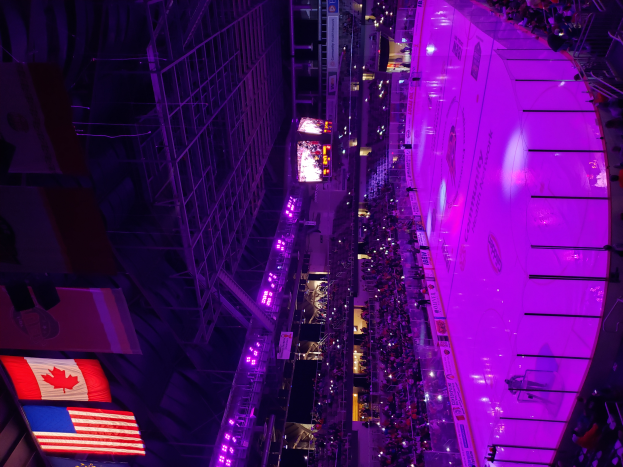 Eine große Menschenmenge steht vor einer hell erleuchteten Bühne, umgeben von Texttafeln und Fahnen, mit einem Gebäude auf der rechten Seite und Eisengittern auf der linken Seite, voller Erwartung auf ein Eishockeyspiel.