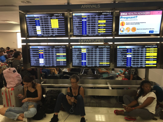 Menschen sitzen auf einem Flughafenboden, einige halten Handys, mit Taschen drumherum und Bildschirmen im Hintergrund, die Zika-Virus-Informationen anzeigen.