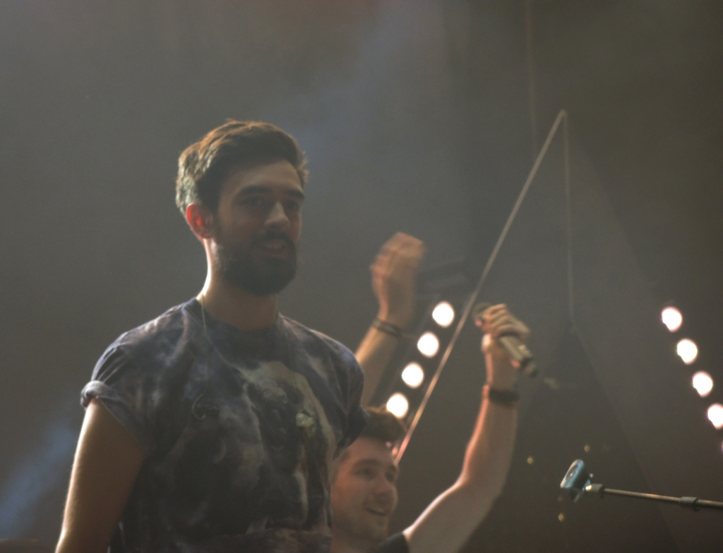 Ein Mann in einem T-Shirt steht und lacht, mit einem anderen lachenden Mann, Lampen, Rauch, einem Mikrofon und anderem Equipment im Hintergrund.