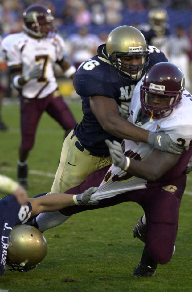 Zwei Fußballspieler in Helmen und Uniformen tackeln sich gegenseitig auf dem Feld, während andere im Hintergrund zusehen.