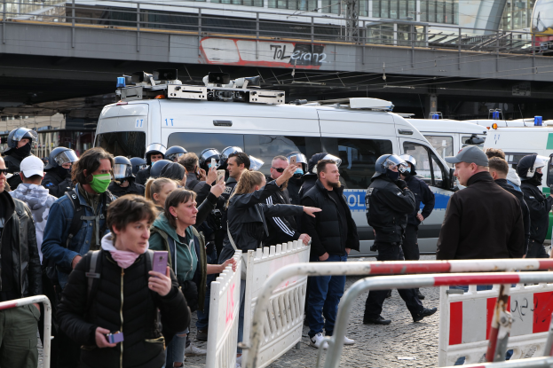 Eine Gruppe von Demonstranten steht hinter Barrikaden und schaut auf Polizeifahrzeuge in Berlin, einige tragen Helme und halten Telefone.