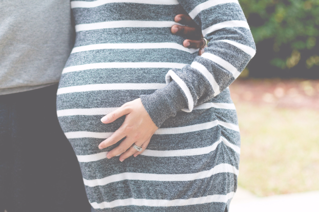 Schwangeres Paar in Umstandskleidung, das zusammen steht und die Hände auf den Bauch legt, umgeben von Bäumen im Hintergrund.