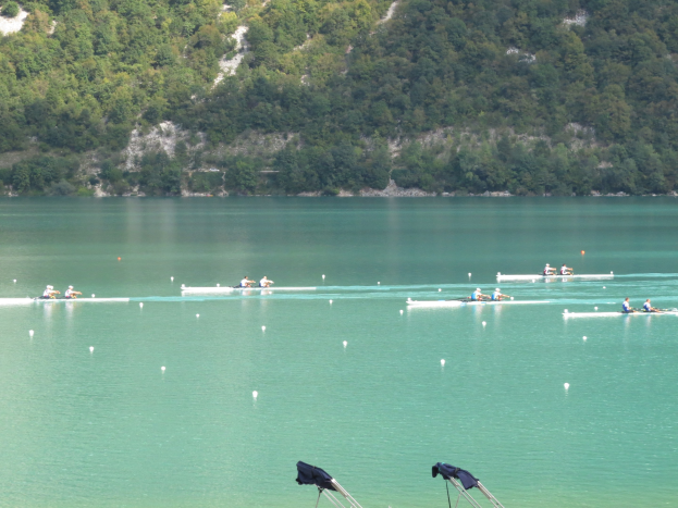 Eine Gruppe von Menschen, die bei einem Wettbewerb auf einem Gewässer mit Bäumen und einem Hügel im Hintergrund rudern, mit Kleidung über den Ständen am Boden.
