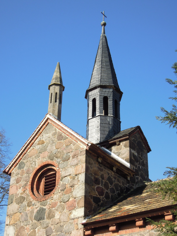 Alte steinerne Kirche mit Turm und Kreuz, umgeben von Bäumen unter einem klaren blauen Himmel.