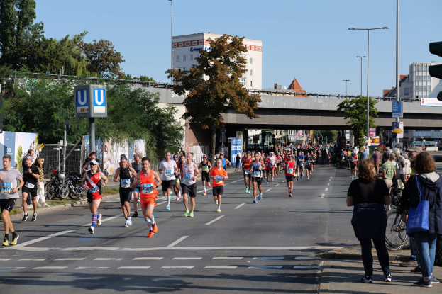 Gruppe von Menschen, die bei einem Marathon auf einer Straße mit Bäumen, Laternen, Schildern, Fahrrädern, einem Zaun, Gras, einer Brücke, Gebäuden und einem klaren blauen Himmel laufen.
