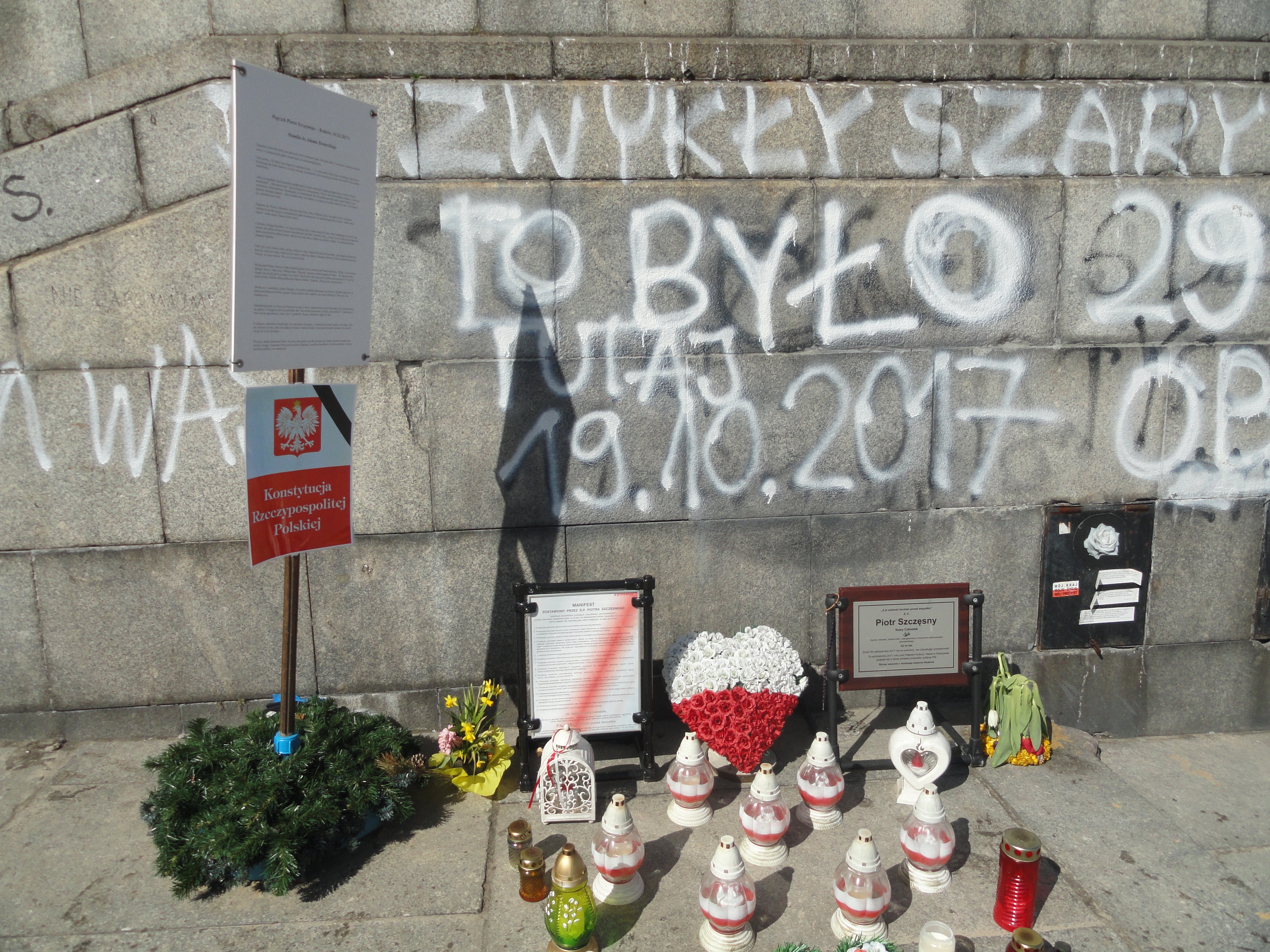 Ein Denkmal mit Blumenstr"{a}ussen, Kerzen und anderen Gegenst"{a}nden vor einer steinernen Wand mit Text und Graffiti, wahrscheinlich im Gedenken an Holocaust-Opfer.