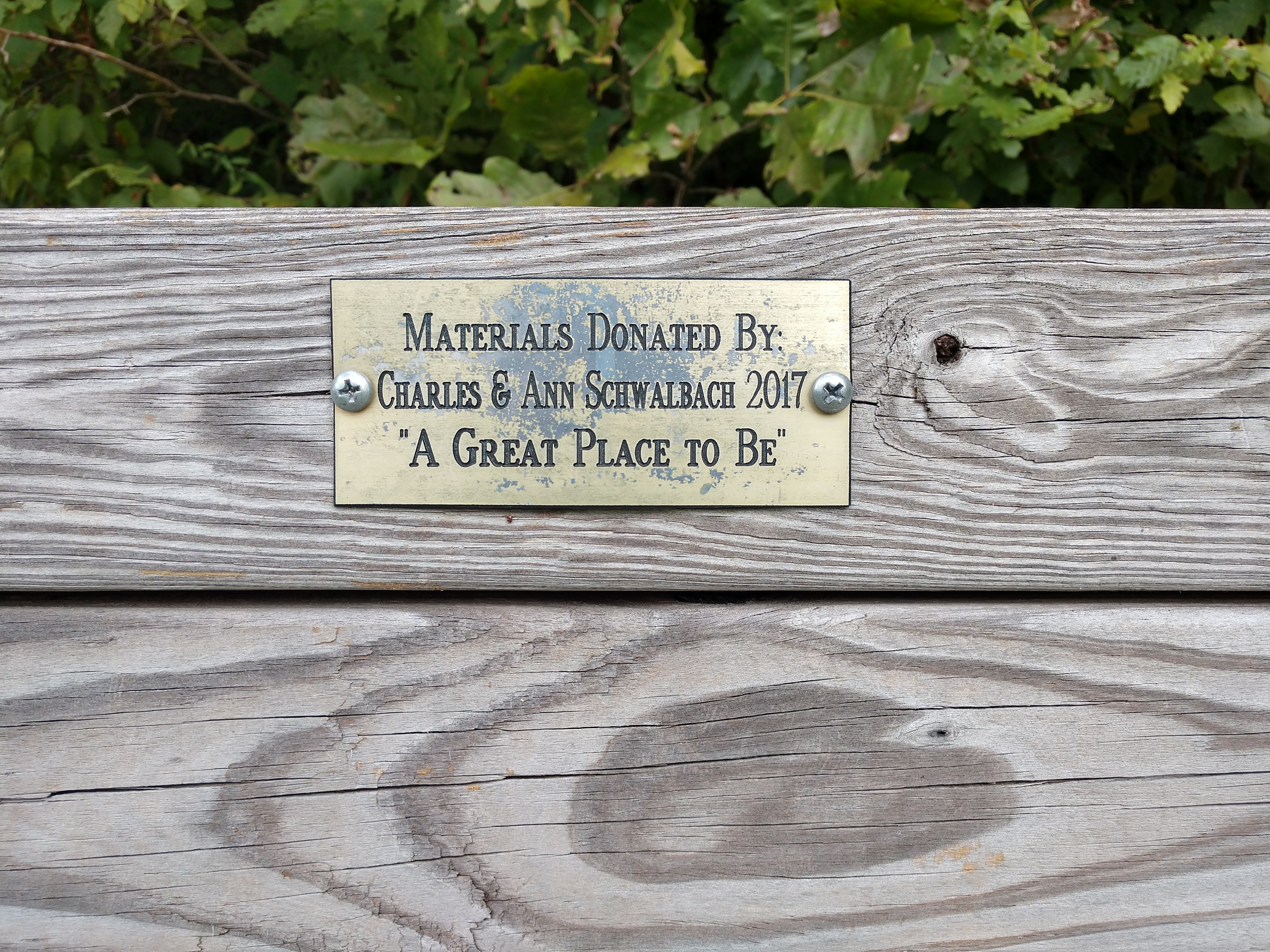 Holzbank mit einer Plakette, auf der "Materialien gespendet von Charles & Ann Schwabach 2017: Ein toller Ort" steht, vor üppigen grünen Pflanzen.