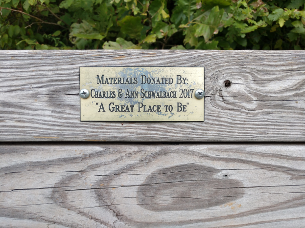 Holzbank mit einer Plakette, auf der "Materialien gespendet von Charles & Ann Schwabach 2017: Ein toller Ort" steht, vor üppigen grünen Pflanzen.