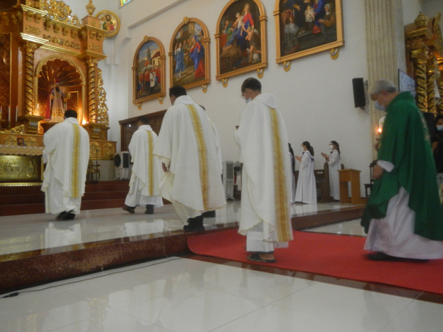 Eine Gruppe von Priestern geht eine rote Teppich in einer Kirche hinunter, mit Menschen, die auf dem Boden vor und rechts von ihnen stehen, und Fotorahmen an der Wand im Hintergrund.