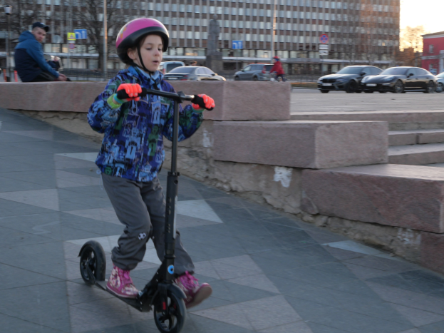 Ein junger Junge in Helm und Handschuhen fährt mit einem Roller eine Treppe hinunter, mit Fahrzeugen, Menschen, Bäumen, Pfosten, Brettern, Gebäuden und einem klaren blauen Himmel im Hintergrund.