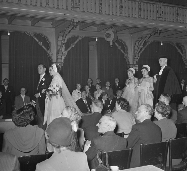 Ein Schwarz-Weiß-Foto einer Hochzeitszeremonie in einem großen Raum mit vielen Gästen, einige halten Blumensträuße, und Tische mit Tassen und anderen Gegenständen davor, mit Vorhängen und Deckenlampen im Hintergrund.