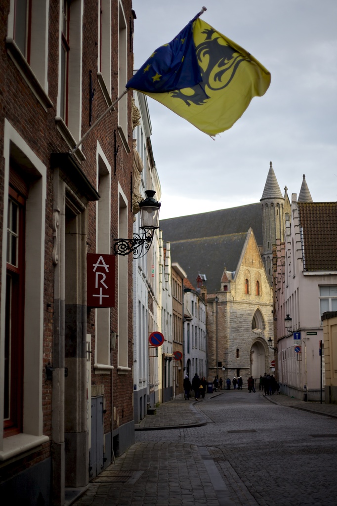Eine Stadtansicht mit Gebäuden, Menschen auf der Straße, Pfählen, Schildern und einer Flagge auf einem der Gebäude, mit dem Himmel oben und der Straße unten.