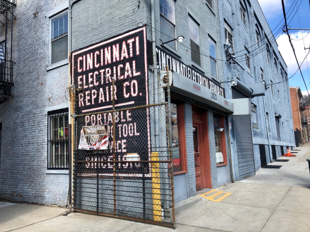 Außenansicht des Cincinnati Electrical Repair Co. Gebäudes mit Schilder, Metallzaun, Mülltonne, Pfählen, Drähten, Bäumen und einem bewölkten Himmel.