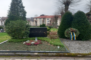 Ein Mahnmal für die Opfer des Zweiten Weltkriegs steht in einem Park, geschmückt mit einem Kranz und umgeben von Grün, mit Gebäuden im Hintergrund.