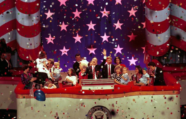 Eine Gruppe von Menschen auf einer Bühne mit einem Podium, das ein Logo, Luftballons und Konfetti zeigt, mit Sternen und einem Vorhang im Hintergrund.