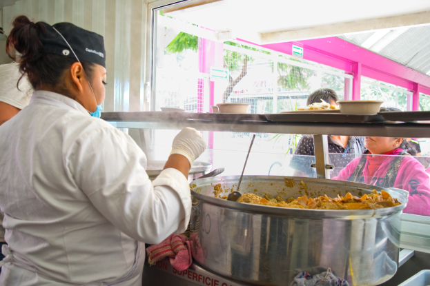 Eine Frau in Kochuniform, weißem Kleid, Handschuhen und schwarzer Mütze, hält einen Löffel und steht vor einer Stahlschüssel mit Essen, mit einer anderen Frau in blauem Kleid links von ihr und verschiedenen Küchenutensilien und Menschen im Hintergrund.