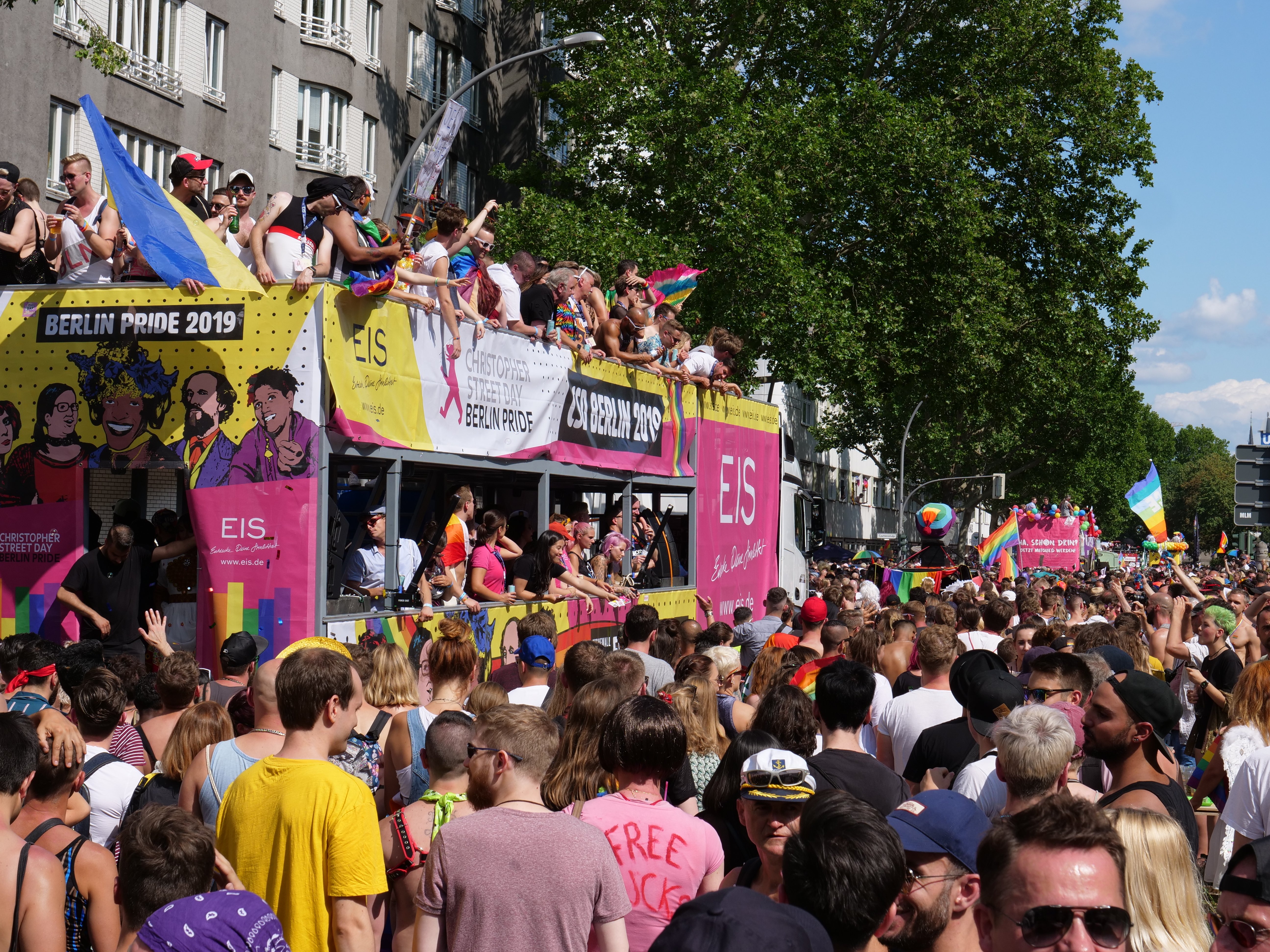 Eine große Menge Menschen, die auf der Straße bei der Berlin Pride 2019 laufen, tragen Kappen und Göggel, halten Fahnen, mit Bannern und Gebäuden im Hintergrund bei einem bewölktem Himmel.