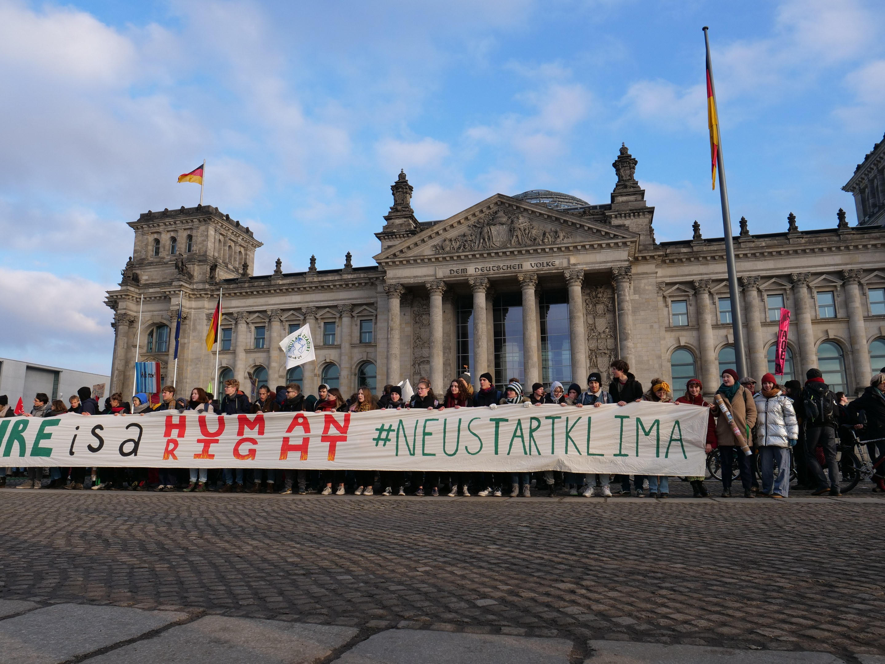 Gruppe von Menschen vor dem Reichstaggebäude in Berlin mit einer Fahne, auf der 'Wir sind ein Menschenrecht' steht, mit den architektonischen Details des Gebäudes und Flaggen im Hintergrund bei bewölktem Himmel.