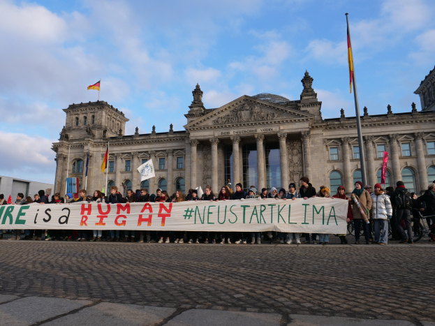 Gruppe von Menschen vor dem Reichstaggebäude in Berlin mit einer Fahne, auf der 'Wir sind ein Menschenrecht' steht, mit den architektonischen Details des Gebäudes und Flaggen im Hintergrund bei bewölktem Himmel.