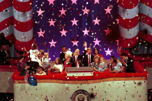 Eine Gruppe von Menschen auf einer Bühne mit einem Podium, das ein Logo, Luftballons und Konfetti zeigt, mit Sternen und einem Vorhang im Hintergrund.