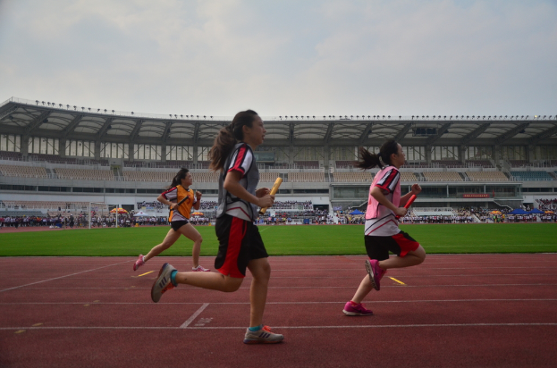 Drei Frauen auf dem Boden laufen und halten Gegenstände, mit Gruppen von Menschen auf einem Weg dahinter und einem Stadion mit Himmel im Hintergrund.